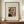 May include: Framed art print featuring Henri Rousseau's 1903 self-portrait showing a balding man with dark hair and mustache wearing formal black attire, displayed on a wooden credenza beside a white ceramic vase and stacked books against white paneled walls