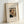 May include: Framed art print of Henri Rousseau's self-portrait showing a man with dark hair, mustache, and formal black suit with white shirt and bow tie, holding a lamp. The vintage-style poster has cream background with elegant typography reading "Self-Portrait of the Artist with a Lamp" and artist signature. Natural wood frame sits propped against white wall on light surface.