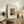 May include: Framed art print displaying Henri Rousseau's self-portrait featuring a balding man with mustache in formal attire, styled as a vintage museum poster with cream matting and thin wooden frame, displayed on rustic wooden sideboard alongside fern plant, books, ceramic vase and table lamp in neutral-toned living room setting