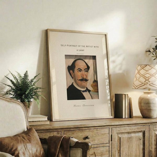 May include: Framed art print displaying Henri Rousseau's self-portrait featuring a balding man with mustache in formal attire, styled as a vintage museum poster with cream matting and thin wooden frame, displayed on rustic wooden sideboard alongside fern plant, books, ceramic vase and table lamp in neutral-toned living room setting