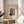 May include: Framed art print of Henri Rousseau's self-portrait showing a man with dark hair, mustache, and formal black suit with bow tie, displayed on a light wood round side table next to a beige upholstered chair. Soft natural light creates shadows across white walls, with a plant visible in the corner. The print features cream matting and thin gold frame.