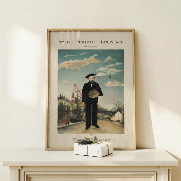 May include: Framed art print of Henri Rousseau's self-portrait showing a bearded man in dark suit holding palette, standing before industrial landscape with bridge and buildings under cloudy sky. Light wood frame displayed on white wooden surface beside small wrapped gift with twine and greenery sprig against cream wall.