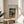 May include: Framed art print featuring Henri Rousseau's painting of a man in dark suit and hat standing near a small child in white dress within a lush green park setting. The poster has cream matting with black text reading "STROLLER AND CHILD" and museum details. Displayed in thin gold frame on round wooden side table next to white upholstered chair, with plant leaves casting shadows on neutral wall in bright natural light.