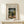 May include: Framed art print featuring Henri Rousseau's naive style painting of an elderly man in dark suit and hat standing beside a small child in white dress on a tree-lined path. The poster has cream matting with black text reading "Stroller and Child" and artist information. Natural wood frame sits on light wooden surface next to small white wrapped gift with twine and greenery sprig against neutral beige wall.