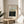 May include: Framed art print featuring a forest landscape with bare trees in muted greens and browns, displayed on a round wooden side table next to a beige upholstered chair. Natural lighting creates soft shadows across cream-colored walls, with a plant leaf visible in the corner of the bright, minimalist room setting.