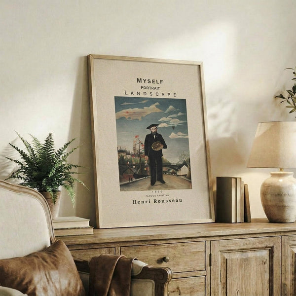 May include: Framed art print featuring Henri Rousseau's self-portrait showing a bearded man in black coat holding a palette, standing before a landscape with cloudy sky and buildings. Print has cream background with black text above and below the artwork. Frame appears light wood, displayed on rustic wooden sideboard alongside fern plant, books, ceramic vase, and lamp in neutral-toned living room setting.