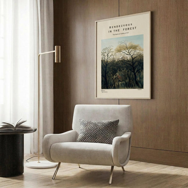 May include: Beige framed poster with forest scene artwork featuring tall trees and figures, displayed on wood panel wall above modern cream armchair with geometric pillow, beside brass floor lamp and dark round side table with open book