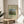 May include: Framed art print of Henri Rousseau's pastoral painting featuring cows grazing in green meadow with tall cypress trees and rounded shrubs under blue sky, displayed on light wood round side table next to beige upholstered chair with plant leaves visible in soft natural lighting
