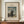 May include: Framed art print of Henri Rousseau's 1890 self-portrait showing a bearded man in black coat holding palette, standing before industrial landscape with bridge and buildings under cloudy sky. Cream matting with brown wooden frame displayed on walnut credenza beside white ceramic vase and stacked books against neutral wall with white trim molding.