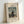 May include: Framed vintage art print featuring a bearded man in dark suit holding a palette, standing in a whimsical landscape with clouds, buildings, and a bridge. The print has cream background with black text reading "MYSELF PORTRAIT LANDSCAPE" at top and "1890 FAMOUS PAINTING Henri Rousseau" at bottom. Light wooden frame sits on white surface against white wall with soft shadows.