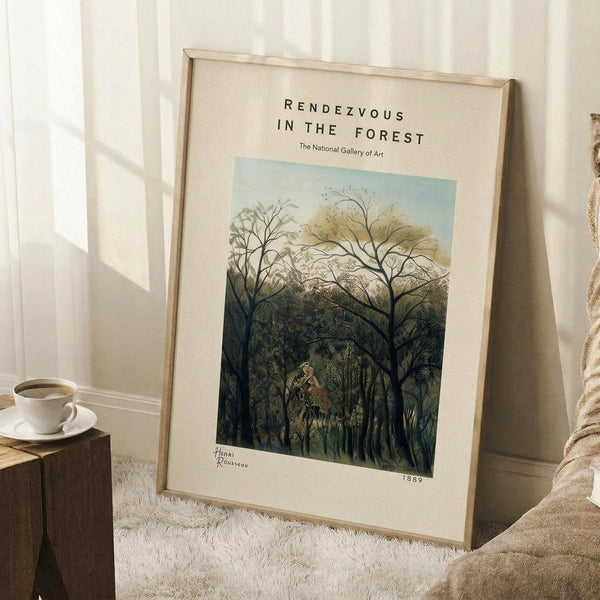 May include: Framed art print leaning against white wall on textured cream rug, featuring lush green forest scene with tall trees and small figures. Natural wood frame, cream matting with museum-style text. Wooden side table with white coffee cup nearby, beige throw blanket visible. Soft natural lighting creates shadows on wall.