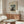 May include: A framed vintage art poster featuring Henri Rousseau's self-portrait showing a bearded man in dark clothing holding a palette, standing before a landscape with ships and buildings. The cream-colored print with black text hangs above a wooden sideboard in a minimalist living room with white curved chair, glass oval coffee table, and modern lamp.