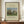 May include: Framed poster print featuring Henri Rousseau's 1910 painting "Meadowland (The Pasture)" with pastoral scene showing cows grazing in green field surrounded by tall cypress trees and rounded foliage under blue sky. Displayed on wooden sideboard next to white ceramic vase and stacked books against cream walls with white trim molding.