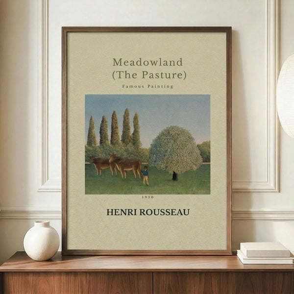 May include: Framed poster print featuring Henri Rousseau's 1910 painting "Meadowland (The Pasture)" with pastoral scene showing cows grazing in green field surrounded by tall cypress trees and rounded foliage under blue sky. Displayed on wooden sideboard next to white ceramic vase and stacked books against cream walls with white trim molding.