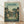 May include: Framed poster print in light wood frame leaning against white wall on wooden floor. Artwork shows Studio Ghibli's My Neighbor Totoro in vintage travel poster style with muted green and yellow tones. Scene depicts traditional Japanese village street at dusk with two children and Totoro character standing under power lines between wooden houses with glowing windows and lush trees.