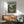 May include: Framed My Neighbor Totoro movie poster displayed above a mid-century modern wood credenza in a sunlit room. The vintage-style poster shows a Japanese village street scene with traditional houses and small figures, featuring muted green and brown tones. A mint green upholstered armchair with wooden frame sits nearby alongside a white ceramic table lamp and round wooden side table.