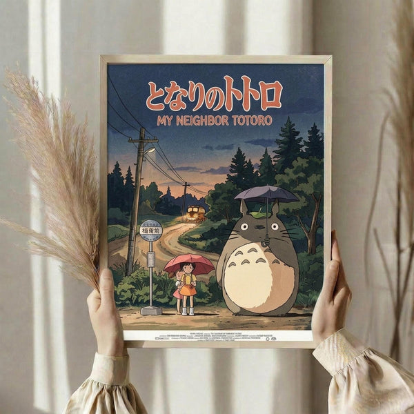 May include: Person holding framed poster featuring Totoro and young girl with red umbrella standing at rural bus stop at dusk. Anime-style artwork shows power lines, forest, and twilight sky in blues and warm tones. Dried wheat stems visible on left side of neutral interior setting.