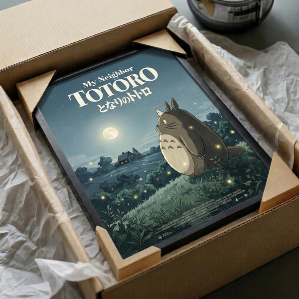 May include: Framed My Neighbor Totoro poster being unboxed from brown cardboard shipping package with white tissue paper. The vintage-style print shows the iconic gray forest spirit character standing in a moonlit landscape with glowing fireflies, rendered in muted blue and green tones. Black frame sits in open cardboard box on white surface next to partial view of dark bowl.