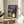 May include: Framed poster featuring iconic anime characters Totoro and a young girl with umbrellas at a rural bus stop, displayed on a round wooden side table next to a beige upholstered chair. The artwork shows a twilight forest scene with power lines, rendered in Studio Ghibli's distinctive animation style with Japanese and English text at the top. Natural lighting creates soft shadows across the minimalist room setting with white walls and a potted plant visible in the corner.