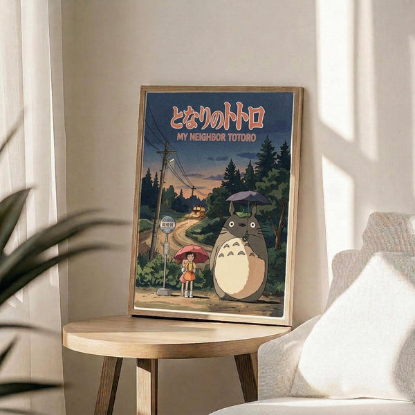 May include: Framed poster featuring iconic anime characters Totoro and a young girl with umbrellas at a rural bus stop, displayed on a round wooden side table next to a beige upholstered chair. The artwork shows a twilight forest scene with power lines, rendered in Studio Ghibli's distinctive animation style with Japanese and English text at the top. Natural lighting creates soft shadows across the minimalist room setting with white walls and a potted plant visible in the corner.