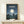 May include: Framed Studio Ghibli movie poster showing nighttime scene with full moon, starry sky, silhouettes of Totoro and children on rolling hills. Dark blue and teal tones with glowing white moon. Japanese text at top with English subtitle. Thin wooden frame displayed on light wood shelf against white wall, small wrapped gift package in front.