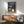 May include: Framed My Neighbor Totoro movie poster displayed above mid-century modern wooden credenza in bright living room setting. Poster shows iconic gray Totoro character standing beside young girl with red umbrella at rural bus stop, surrounded by green forest and telephone poles at dusk. Warm wood frame complements the teal upholstered armchair and ceramic table lamp nearby, with natural sunlight creating geometric shadows on white wall.