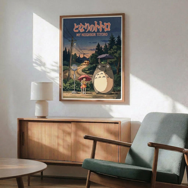 May include: Framed My Neighbor Totoro movie poster displayed above mid-century modern wooden credenza in bright living room setting. Poster shows iconic gray Totoro character standing beside young girl with red umbrella at rural bus stop, surrounded by green forest and telephone poles at dusk. Warm wood frame complements the teal upholstered armchair and ceramic table lamp nearby, with natural sunlight creating geometric shadows on white wall.