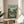 May include: Framed poster featuring an illustrated Japanese village street scene with traditional houses, power lines, and small figures walking with an umbrella. The artwork displays muted green and earth tones in an anime-inspired style. The wooden-framed print sits on a light wood round side table next to a beige upholstered chair and plant, with natural window light casting shadows across white walls.