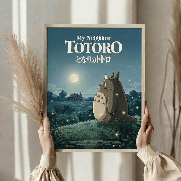 May include: Person in cream sweater holding framed poster against white curtained window, with dried pampas grass. Poster shows large gray forest creature sitting in moonlit field with fireflies, village silhouette in background. Blue evening sky with full moon and Japanese text below English title. Light wooden frame.