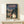 May include: Framed poster displaying Studio Ghibli's iconic Totoro character standing at a rural bus stop with a young girl holding a red umbrella, set against a twilight forest scene with power lines and a distant bus. The vintage-style artwork features muted blues, greens, and warm tones in a wooden frame, positioned on a light wood shelf beside a small wrapped gift against a cream wall.