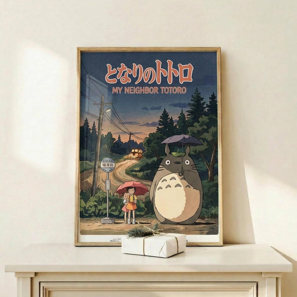 May include: Framed poster displaying Studio Ghibli's iconic Totoro character standing at a rural bus stop with a young girl holding a red umbrella, set against a twilight forest scene with power lines and a distant bus. The vintage-style artwork features muted blues, greens, and warm tones in a wooden frame, positioned on a light wood shelf beside a small wrapped gift against a cream wall.
