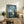 May include: Framed poster displayed on rustic wooden sideboard in cozy living room setting. Features iconic gray forest creature character standing in moonlit field with small glowing spirits, house silhouette in distance. Blue-toned nighttime scene with Japanese text below English title. Styled with fern plant, ceramic vase, books, and beige armchair with brown throw blanket.