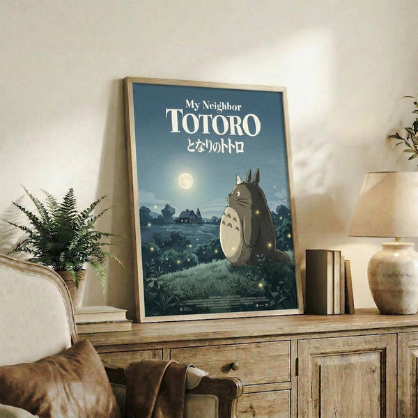 May include: Framed poster displayed on rustic wooden sideboard in cozy living room setting. Features iconic gray forest creature character standing in moonlit field with small glowing spirits, house silhouette in distance. Blue-toned nighttime scene with Japanese text below English title. Styled with fern plant, ceramic vase, books, and beige armchair with brown throw blanket.