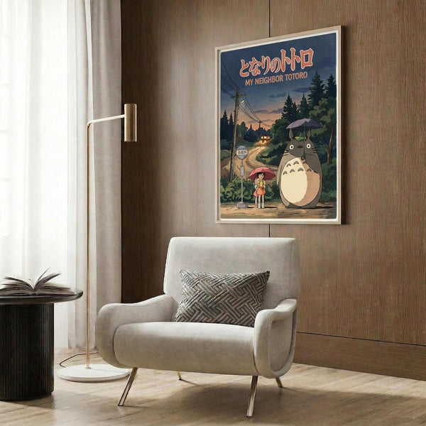May include: Framed My Neighbor Totoro poster hanging on wood-paneled wall above beige upholstered armchair with geometric pillow. The vintage-style poster shows Totoro and a child with umbrella at a bus stop surrounded by forest scenery in muted blue and green tones. Modern living room setting with gold floor lamp and side table.
