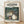 May include: Framed poster displaying Studio Ghibli's My Neighbor Totoro movie artwork in natural wood frame, leaning against white wall on light wooden floor. Features iconic gray Totoro character holding umbrella next to two young girls at forest bus stop, with large cat-shaped bus in background among lush green trees. Japanese and English text at top, vintage film poster style with muted earth tones and whimsical animation aesthetic.
