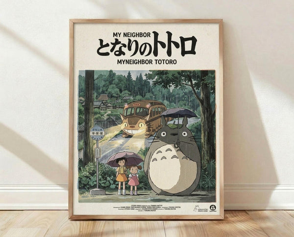 May include: Framed poster displaying Studio Ghibli's My Neighbor Totoro movie artwork in natural wood frame, leaning against white wall on light wooden floor. Features iconic gray Totoro character holding umbrella next to two young girls at forest bus stop, with large cat-shaped bus in background among lush green trees. Japanese and English text at top, vintage film poster style with muted earth tones and whimsical animation aesthetic.