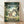 May include: Framed poster of Studio Ghibli's My Neighbor Totoro leaning against white wall on wooden floor. Features large gray Totoro with smaller blue and white Totoro creatures alongside three children in forest setting with traditional Japanese house. Vibrant greens and earth tones in anime art style. Natural wood frame, soft lighting creates gentle shadows on textured white wall surface.