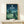 May include: Framed Studio Ghibli My Neighbor Totoro movie poster displayed on light wood shelf with small wrapped gift box. Poster shows nighttime scene with crescent moon, stars, constellations, large gray Totoro character with two children in grassy meadow surrounded by lush green trees. Japanese text at top with English translation below. Artwork features deep blues and greens in whimsical animation style with glowing fireflies scattered throughout scene.