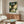 May include: My Neighbor Totoro movie poster displayed above a mid-century modern walnut credenza in a minimalist living room. The vintage-style poster shows Totoro and characters under a large tree in a magical forest setting with Japanese text. Room features a curved white chair, glass-top oval coffee table, and warm wood tones throughout.