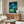 May include: Studio Ghibli My Neighbor Totoro movie poster displayed on white wall above wooden sideboard in modern living room. Poster features nighttime forest scene with children, large gray Totoro character, crescent moon and stars in deep blue-green tones. Room includes cream upholstered chair, glass-top coffee table, and minimalist decor with natural wood finishes throughout.