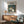 May include: Studio Ghibli My Neighbor Totoro movie poster in wooden frame hanging on white wall above mid-century modern wooden credenza. Gray upholstered armchair with wooden arms sits nearby. Poster shows iconic gray Totoro character with yellow Catbus and children under umbrellas in forest setting with Japanese text. Warm natural lighting creates soft shadows across the minimalist room scene.