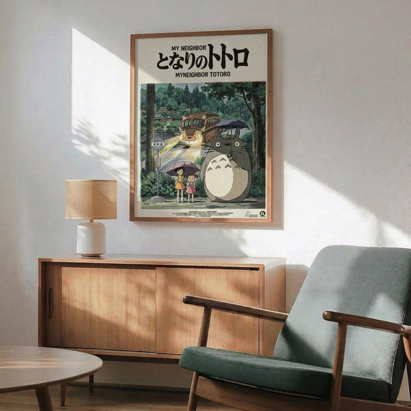 May include: Studio Ghibli My Neighbor Totoro movie poster in wooden frame hanging on white wall above mid-century modern wooden credenza. Gray upholstered armchair with wooden arms sits nearby. Poster shows iconic gray Totoro character with yellow Catbus and children under umbrellas in forest setting with Japanese text. Warm natural lighting creates soft shadows across the minimalist room scene.