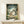 May include: Framed poster displaying animated forest scene with large gray creature, family of three people, and small white creatures among green trees. Wooden frame sits on light wooden shelf next to small white wrapped package against cream wall.