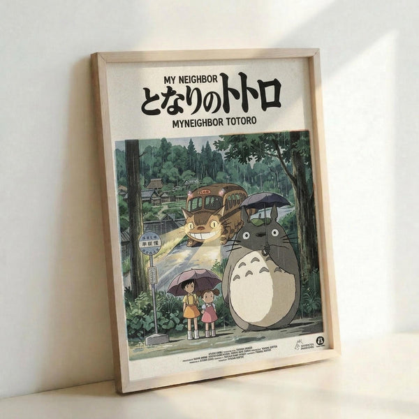 May include: Framed poster print featuring beloved animated characters Totoro and Catbus from Studio Ghibli film, displayed in natural wood frame against white wall. Artwork shows large gray forest spirit holding umbrella with two children at rainy bus stop, cat-shaped bus visible among lush green trees. Japanese text appears at top with English translation. Soft natural lighting creates gentle shadows on wall.