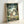 May include: Framed poster print leaning against white wall showing animated forest scene with large gray creature, family of characters, and smaller woodland beings among green trees. Natural wood frame, soft lighting creates gentle shadows on wall surface.