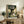 May include: Framed Studio Ghibli movie poster featuring Totoro and children under a large forest tree, displayed on rustic wooden sideboard with fern plant, books, ceramic vase, and beige armchair visible in cozy living room setting with neutral walls