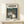May include: Framed Studio Ghibli My Neighbor Totoro movie poster in natural wood frame, displayed on light wooden shelf against white wall. Artwork shows iconic gray Totoro character with two children holding umbrellas at forest bus stop, with Catbus in background among lush green trees. Small white wrapped gift box sits in front of frame. Vintage-style Japanese text at top, warm earth tones throughout illustration.