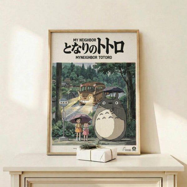 May include: Framed Studio Ghibli My Neighbor Totoro movie poster in natural wood frame, displayed on light wooden shelf against white wall. Artwork shows iconic gray Totoro character with two children holding umbrellas at forest bus stop, with Catbus in background among lush green trees. Small white wrapped gift box sits in front of frame. Vintage-style Japanese text at top, warm earth tones throughout illustration.