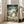 May include: Large framed poster of animated forest scene with large gray creature, three children, and smaller woodland beings among green trees and wooden structures. White or light wood frame leans against beige wall near cream-colored armchair in bright living room with white trim and windows.