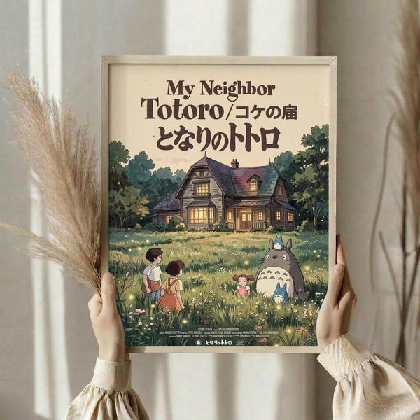 May include: Hands holding a framed poster showing an illustrated scene with two children, large gray creature Totoro, and smaller woodland spirits in a grassy meadow. A cozy house with glowing windows sits among green trees in the background. The poster has Japanese and English text at the top. Dried wheat stalks visible on the left side, neutral curtains in soft lighting.